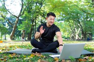 Fitness instructor works out online uses laptop in park sitting in lotus position, Asian athlete communicates with students remotely, call training online remotely photo