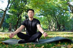 Portrait of Asian man in autumn park meditating sitting in lotus position on fitness mat, sportsman resting after training doing breathing exercises, photo