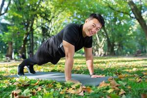 retrato de contento masculino atleta, asiático mirando a cámara y sonriente haciendo físico ejercicios, Lagartijas y tablón en otoño parque en soleado día. foto