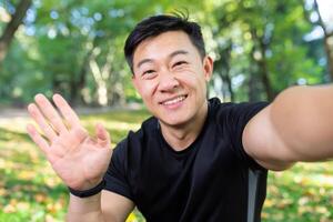 asiático deportista corredor en otoño parque mirando a teléfono inteligente cámara y sonriente, hablando con amigos en llamar, hombre en ropa de deporte para aptitud en otoño bosque. foto