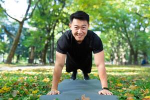 Portrait of happy male athlete, Asian looking at camera and smiling doing physical exercises, push-ups and plank in autumn park on sunny day. photo