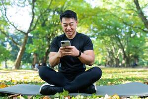 Sportsman in lotus pose sitting on sports mat in park and smiling, asian man using phone online watching and typing message photo