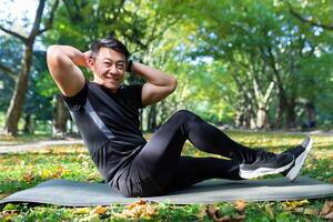 retrato de un atleta en el parque, un asiático hombre mira a el cámara y sonrisas, un hombre en un Deportes traje lo hace físico ejercicios en un Deportes estera. foto
