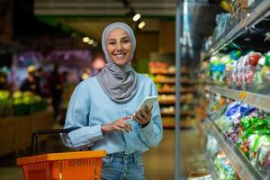 satisfecho árabe mujer en hijab es compras en el almacenar, participación un teléfono y un compras cesta, sonriente y mirando a el cámara. foto