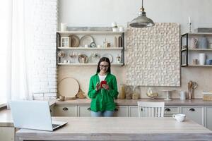 hermosa mujer en lentes y verde camisa utilizando rojo móvil teléfono a hogar en cocina, sonriente y contento foto