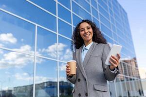 Happy Latin American business woman walks past the glass office building, holds a cup of coffee on the phone, smiles and looks at the camera. photo