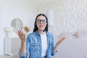 Beautiful woman looking at web camera, nervous and arguing with interlocutor, using headset for call, working at home sitting on sofa, online conference and study photo