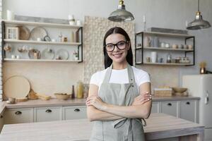 retrato de un joven hermosa mujer en el cocina, un morena vistiendo un gris delantal es sonriente y mirando a el cámara foto