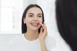 hermosa joven mujer en un blanco camiseta en el Mañana mira en el espejo y sonrisas examinando su dientes y maquillaje foto