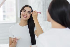 un joven hermosa morena mujer a hogar mira en el espejo en el mañana, sonrisas, aplica maquillaje con un cepillo foto