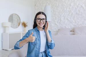 Beautiful woman working and studying at home using headset for call, smiling and looking at camera, holding finger up, sitting on sofa in living room photo