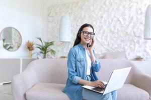 Portrait of young beautiful woman, working and studying online with laptop, using headset for call, working remotely at home, sitting in living room on sofa, happy looking at camera photo