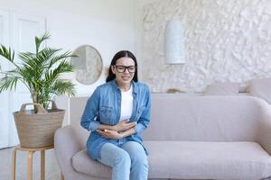 A young beautiful woman sits on the couch at home, sick, has severe pain in her stomach and side photo