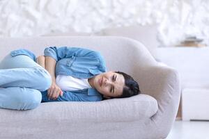 Young beautiful woman alone at home, lying on the sofa, close-up photo, upset, having a severe stomach ache photo