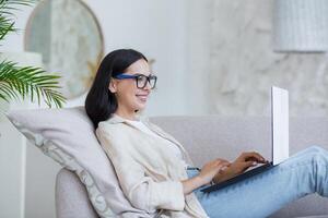 Young beautiful girl studying online at home sitting on sofa in living room, brunette in glasses working with laptop remotely photo