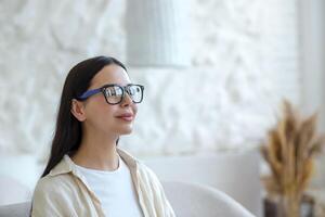 Young beautiful woman in glasses resting with eyes closed, breathing fresh cool air, brunette at home sitting on sofa, dreaming and meditating close-up photo