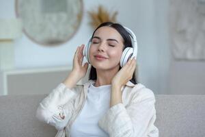 Beautiful young woman resting listening to music in white big headphones, brunette sitting on sofa in living room with closed eyes dreaming at home photo