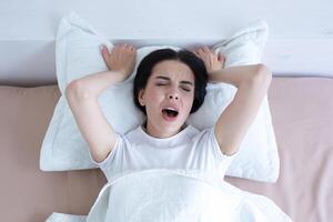 Frustrated and upset woman lying in bed alone at home crying and screaming, top view, brunette in white t-shirt pajamas photo