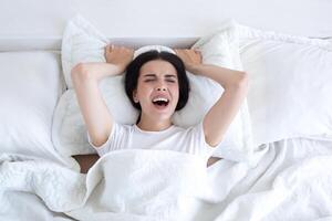 Frustrated and upset woman lying in bed alone at home crying and screaming, top view, brunette in white t-shirt pajamas photo