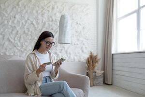 hermosa joven mujer a hogar en lentes sentado en el sofá utilizando un móvil teléfono teléfono inteligente para en línea compras en un almacenar, participación un banco crédito tarjeta en su manos foto