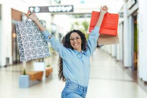 Happy hispanic woman with curly hair in the store looking at the camera and smiling, dancing and happy with a successful shopping, holding colorful bags with goods in her hands, dancing and jumping. photo