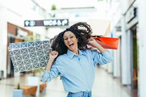 Happy shopper woman looking at camera and smiling, hispanic woman with curly hair dancing and jumping with pleasure, bought gifts on sale, portrait of happy shopping woman. photo