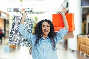 Happy young attractive hispanic woman holding colorful shopping bags in her hands in the shopping center, dancing, rejoicing, jumping, raising the bags up. Looking at the camera, smiling. photo