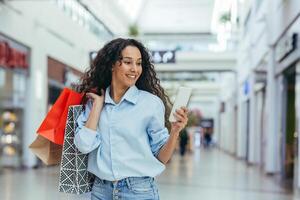 contento mujer compras para ropa durante el período de descuentos y promocional ofertas, Hispano mujer sonriente y contento hojeada en línea descuentos utilizando teléfono inteligente, dentro un moderno grande almacenar. foto