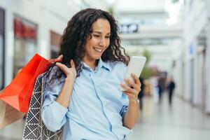 Happy and successful woman shopping for clothes in a supermarket store, Hispanic woman holding a smartphone reading online messages and browsing offers with discounts and sales photo