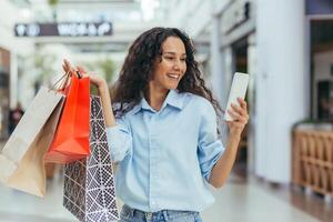 An attractive young Latin American woman is shopping in a shopping center, holding colorful bags in her hands. Uses the phone, takes a selfie, smiles. photo