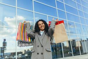 hermosa mujer con Rizado pelo fuera de grande Tienda mirando a cámara y sonriente, Hispano mujer comprador contento con descuentos y venta, en calentar Saco retrato. foto