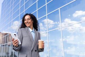 Portrait of a young beautiful Latin American businesswoman in a suit walking outside near a glass office center. Holds a cup of coffee, looks at the phone, smiles. photo
