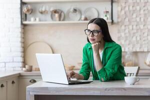 Upset young woman sitting at home in the kitchen in front of a laptop. Type on the keyboard, search, check. Thoughtfully, she rested her head on her hand. photo