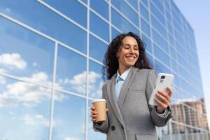 Successful business woman with smartphone goes to work in the morning in the office, hispanic woman with curly hair smiling and browsing online news outside with cup hot drink, in warm clothes coat. photo