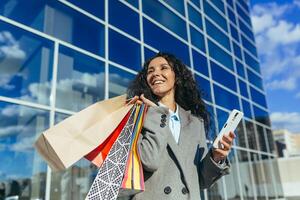 Happy woman shopper outside near big mall store, holding smartphone and colorful packages with goods and gifts, hispanic woman with curly hair in warm coat on sunny day looking away photo