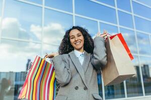 hermosa mujer con Rizado pelo fuera de grande Tienda mirando a cámara y sonriente, Hispano mujer comprador contento con descuentos y venta, en calentar Saco retrato. foto