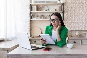 A young woman sits at home in the kitchen with a laptop, jots down the family budget in a notebook, counts bills, keeps a list of purchases. photo