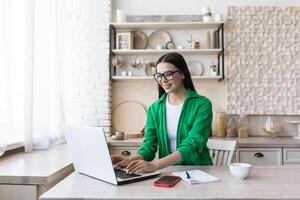 A young woman journalist, writer, blogger works remotely from home using a laptop online. Sitting in the kitchen at the table, making notes in a notebook. photo