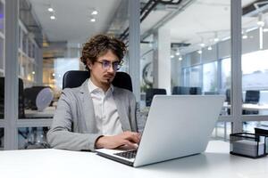 Serious and focused businessman inside modern office working on laptop, bearded man typing on keyboard, thinking man in business clothes sitting at workplace. photo