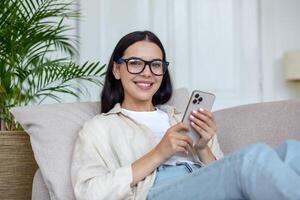 joven hermosa mujer en lentes es acostado en sofá y utilizando móvil teléfono, mirando a cámara, sonriente. foto