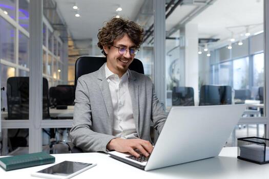 alegre y exitoso empresario dentro oficina trabajando con computadora portátil, barbado hombre sonriente y mecanografía en ordenador portátil teclado, programador codificación software en moderno habitación. foto