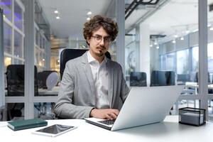 Portrait of thinking serious businessman inside office, bearded man looking into camera focused in glasses, worker in business clothes sitting at workplace using laptop at work. photo