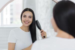 Close-up photo. A beautiful young woman stands and looks at herself in the mirror. She combs her hair with a brush, does her hair. She smiles, looking at the camera. photo