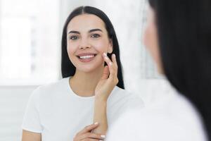 Portrait of a young woman stands and looks at herself in the mirror. She touches her face with her hands, looking at the camera, smilling, photo