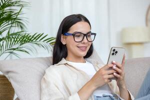 A young beautiful woman is lying on the sofa at home with a phone in her hands. Makes food orders for home, shopping in an online store. photo