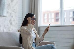 A young woman is sitting at home on the sofa near the window with a phone. plays online games, lottery, raffle. Celebrates the victory, rejoices at the win, shows yes with his hand. photo