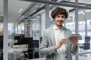 retrato de exitoso joven empresario, hombre con tableta computadora en manos sonriente y mirando a cámara, empresario a lugar de trabajo dentro moderno oficina. foto