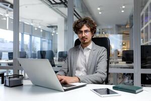 Portrait of thinking serious businessman inside office, bearded man looking into camera focused in glasses, worker in business clothes sitting at workplace using laptop at work. photo