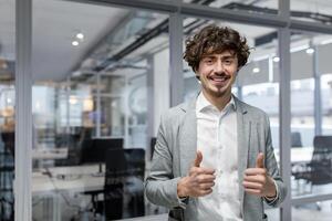 retrato de exitoso empresario jefe, gerente en negocio traje y barba mirando a cámara y sonriente en pie cerca ventana, demostración pulgares arriba, firmar de éxito y lograr metas foto