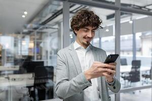 exitoso empresario con barba en pie cerca ventana, hombre utilizando teléfono, contento jefe mecanografía mensaje y hojeada en línea paginas en teléfono inteligente aplicación dentro oficina foto